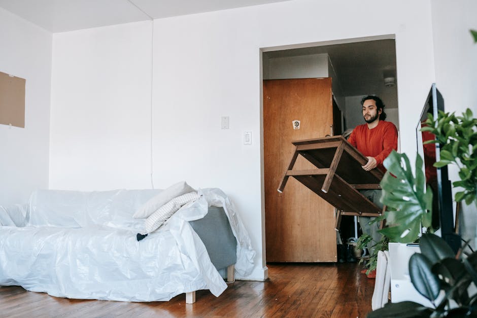 A minimalist interior scene featuring light-colored wooden furniture, including a tall, slatted backrest chair positioned on the right side of the image and a low, rectangular sideboard with rounded e