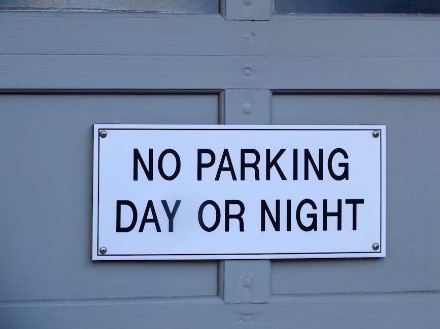 A close-up of a rectangular white parking restriction sign mounted on a metal pole against a bright blue sky with some faint cloud formations. The sign features a black capital letter 'P' with a red c