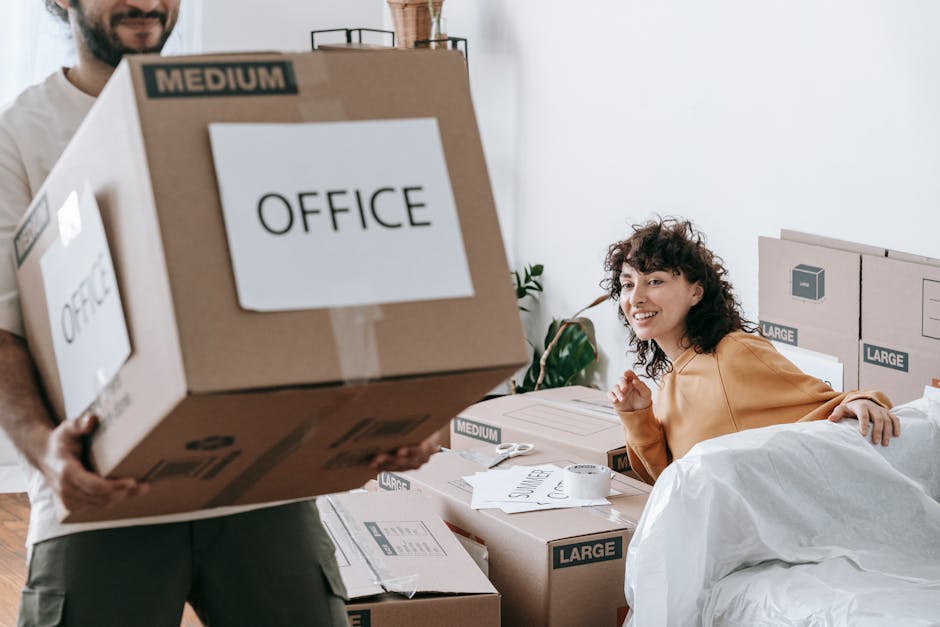 A woman with curly dark hair, wearing a mustard-colored top, is seated on a bed surrounded by unpacked moving boxes labeled 'OFFICE', 'LARGE', and 'MEDIUM'. She is smiling and appears to be engaging with a man, who is partially visible on the left side of the image, holding two large cardboard boxes marked 'OFFICE'. The boxes are made of cardboard, some wrapped in plastic, and are stacked on a wooden surface and on the floor. The scene is set inside a room with white walls, a window allowing natural light, and a small potted plant in the background. Cardboard packing materials, including tape and paper, are scattered on the bed and nearby surfaces, indicating an active home relocation process. The manandavanpimlico.co.uk team is experienced in furniture transport and packing, supporting efficient moving logistics during the house removals service in Pimlico.