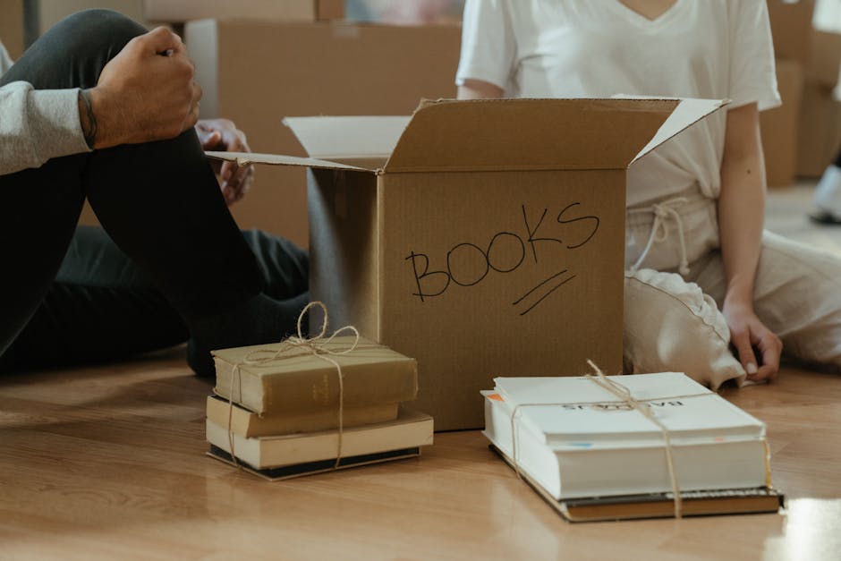 A young woman is sitting on a wooden floor inside a residential space, surrounded by multiple large cardboard boxes used for house removals. She is wearing a white tank top, beige pants, and black-and