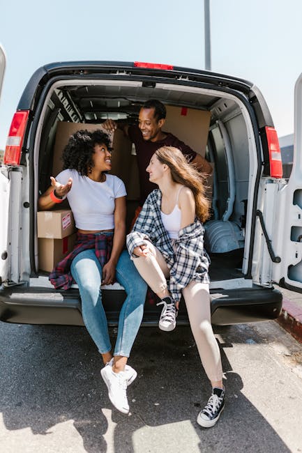 Three individuals are situated at the back of a white cargo van, which is positioned outside a residential property during daylight hours. The van's rear doors are wide open, revealing an interior fil